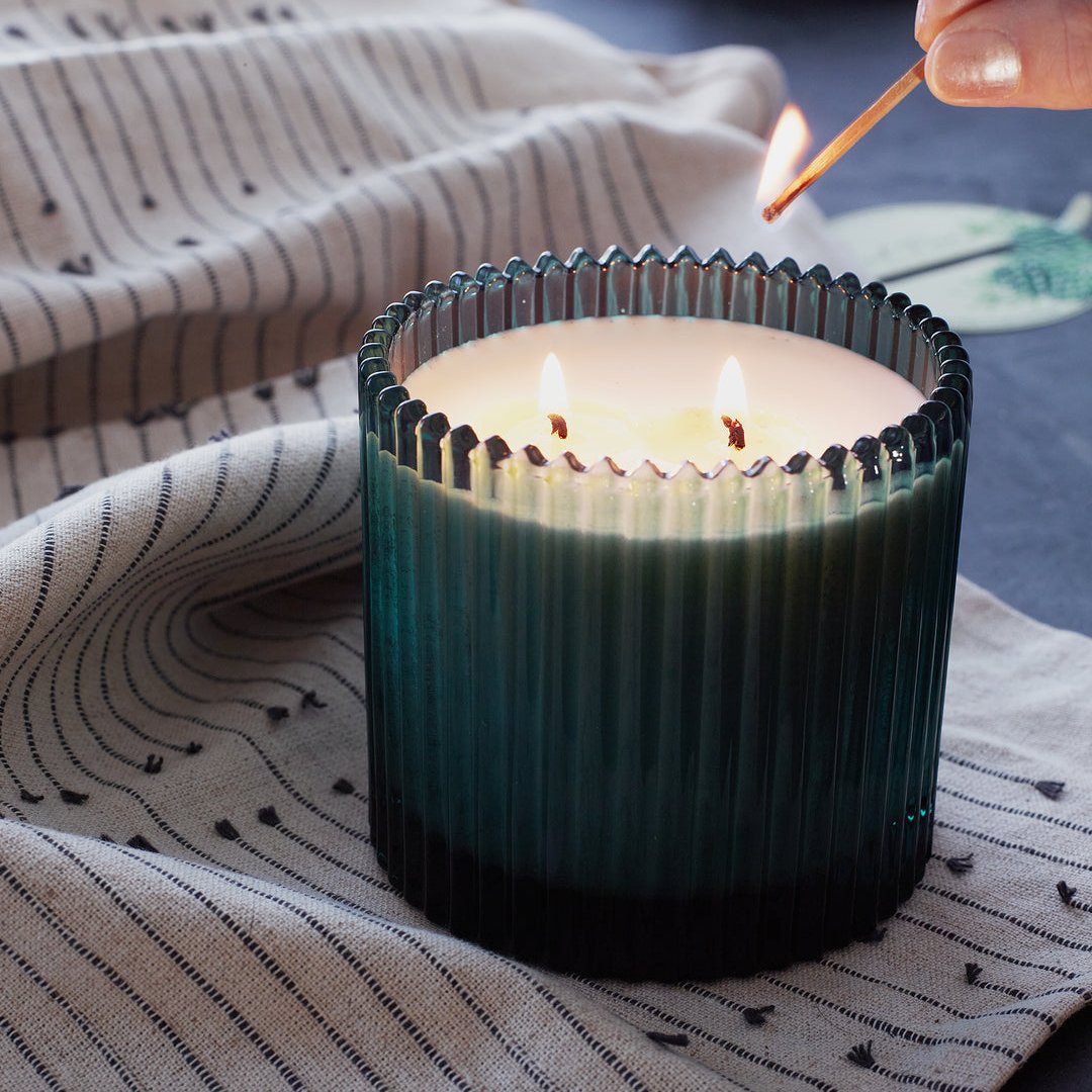 A reaching down and lighting 2-wick green glass candle with a match. Candle is on top of a draped cloth on a flat surface. 