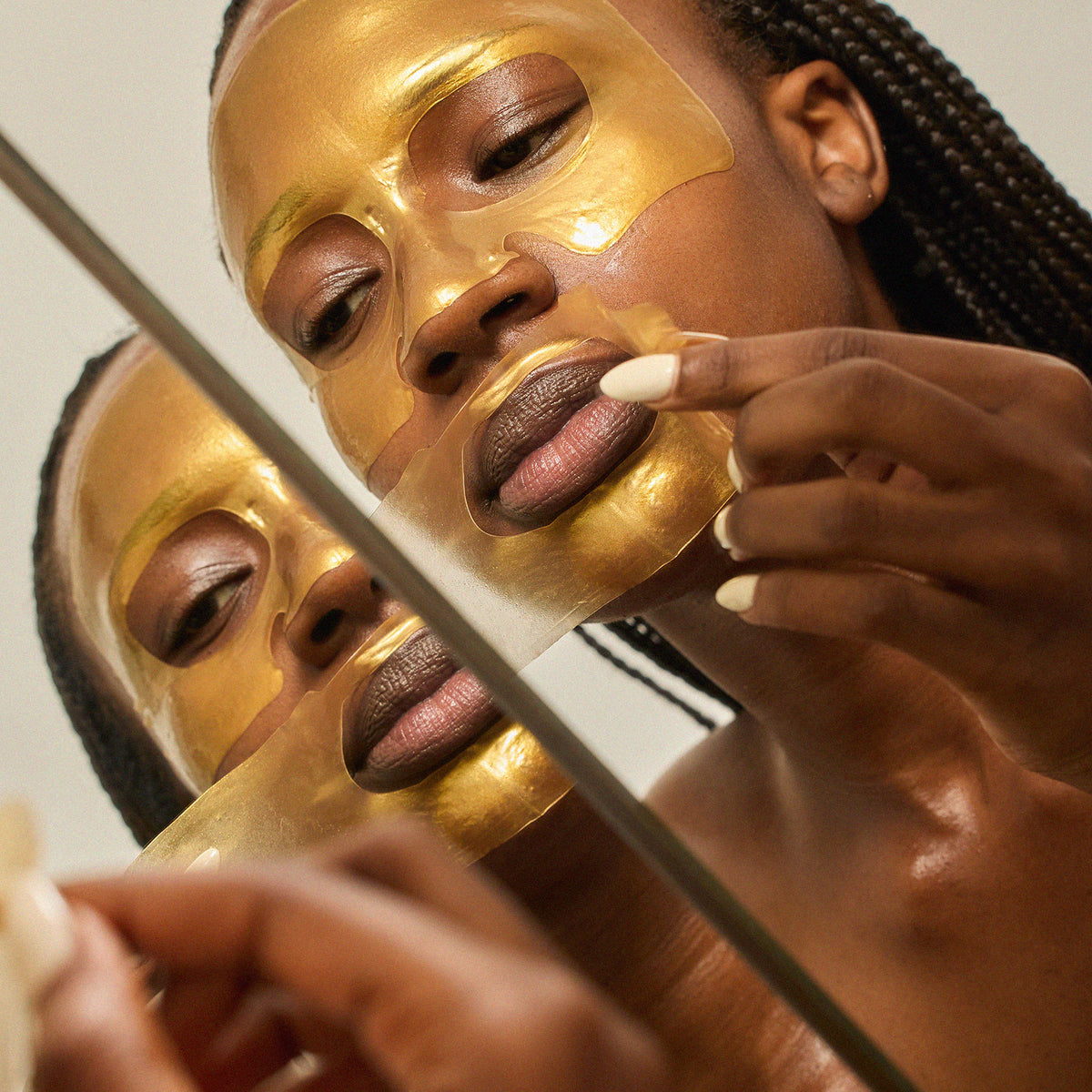 A close-up lifestyle shot of a woman applying Radford Fix -the face mask facial mask, with her reflection visible in a mirror. She has dark braided hair and is wearing a gold mask on her face.