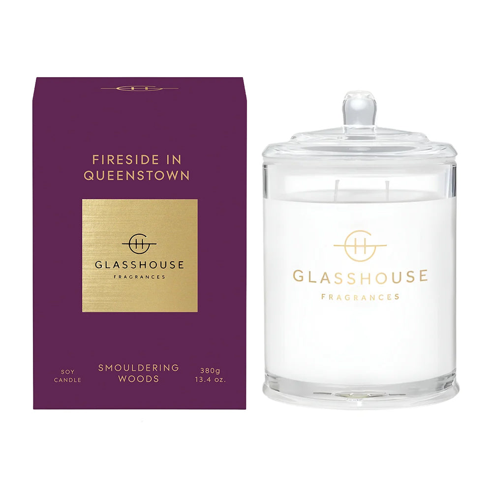 A glass jar candle with white wax and a clear lid stands beside its purple retail box. The box and label feature gold text and a gold square panel, with the brand name GLASSHOUSE FRAGRANCES clearly visible.