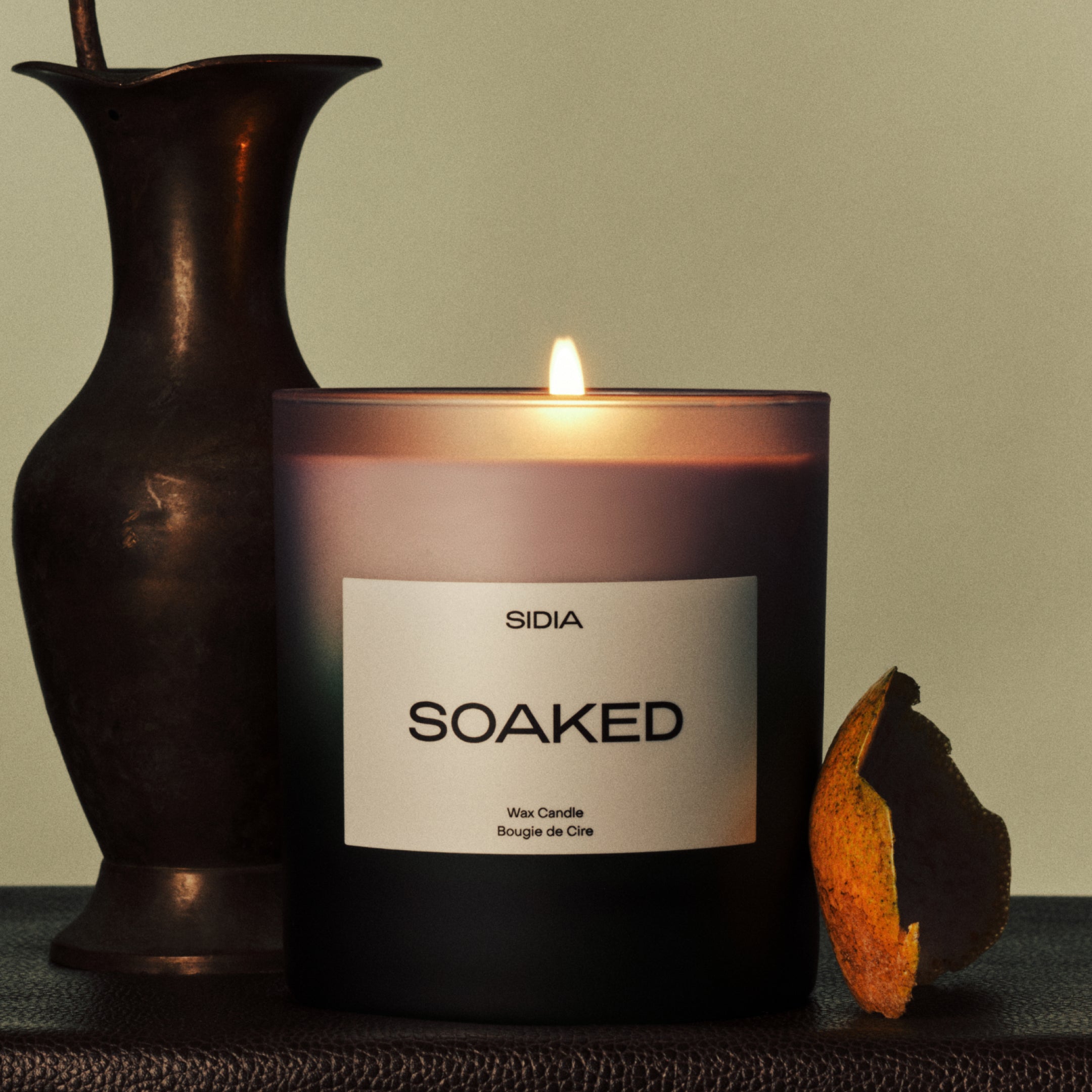 SIDIA candle in "SOAKED" scent leather surface next to an orange peel and a vase