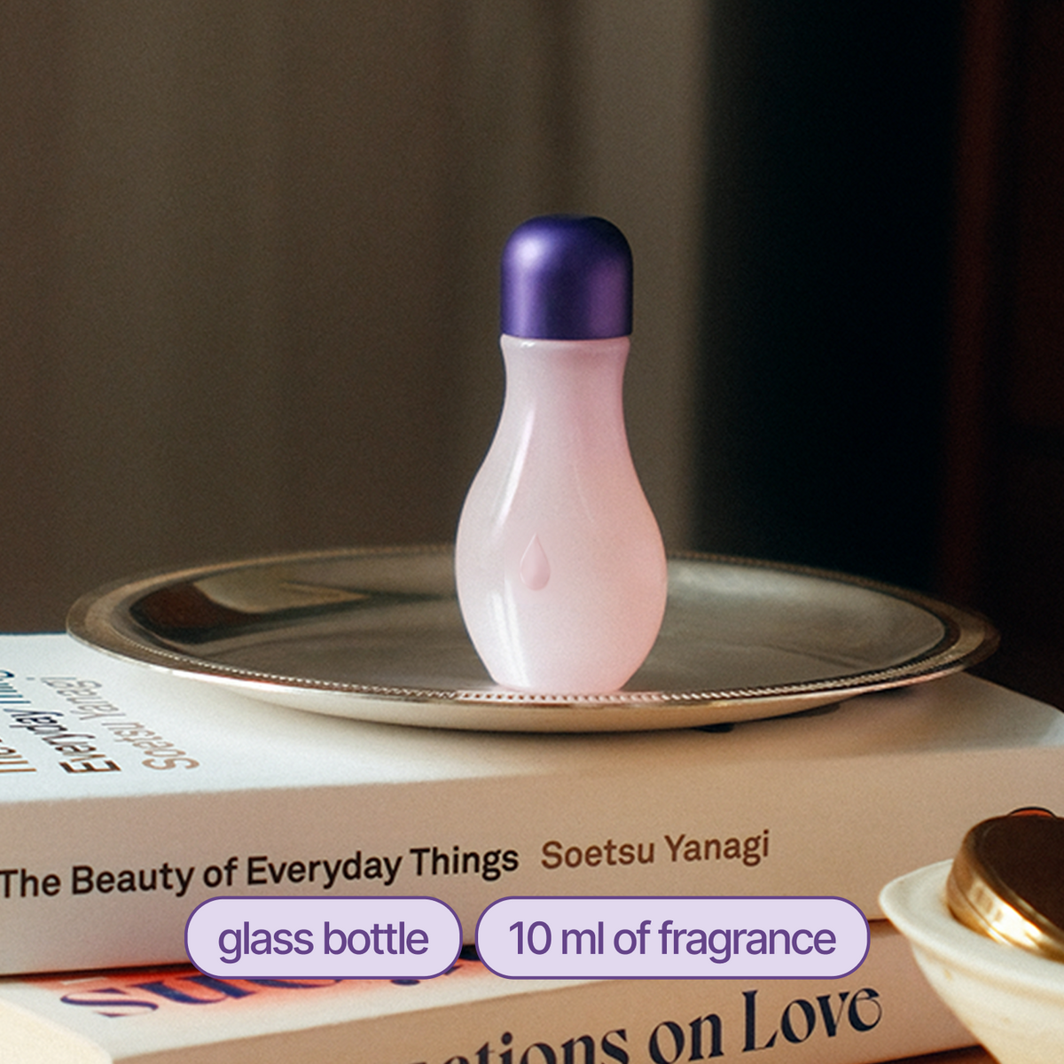 A small glass fragrance bottle with a pale pink body and a metallic purple cap, displayed on a silver tray resting on stacked books.