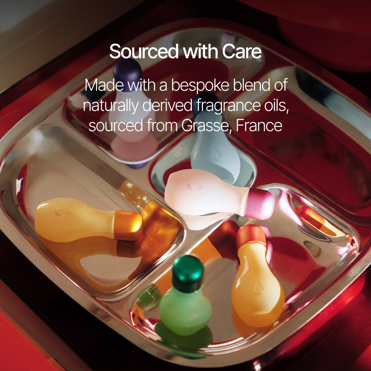 A stainless steel tray holding several small perfume bottles with colorful caps and bodies in amber, pink, green, white, and blue tones. White text overlays read 'Sourced with Care' and 'Made with a bespoke blend of naturally derived fragrance oils, sourced from Grasse, France.