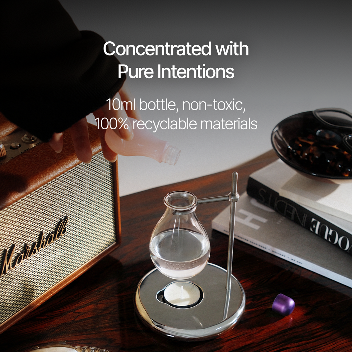 A hand pours liquid from a small bottle into a clear glass vial on a metal stand, set on a wooden table beside a Marshall speaker, books, and a dark bowl. White text reads: 'Concentrated with Pure Intentions', '10ml bottle, non-toxic, 100% recyclable materials'.