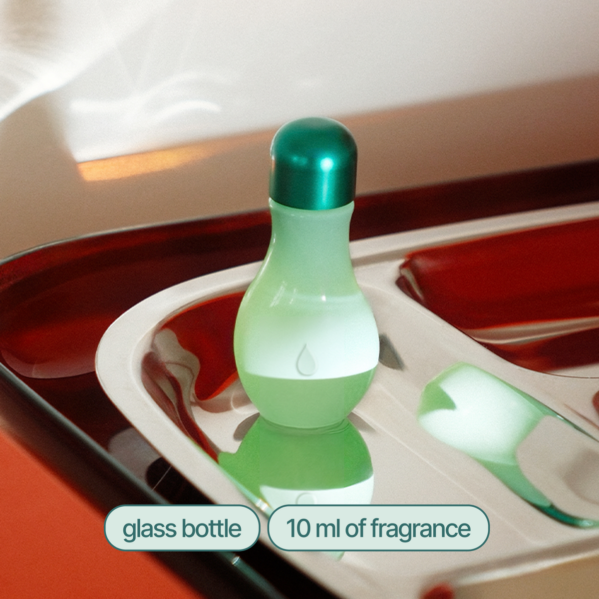 A small glass fragrance bottle with a rounded bulbous body, frosted green to white gradient, and a metallic teal cap, shown on a tray with overlaid text reading 'glass bottle' and '10 ml of fragrance'.