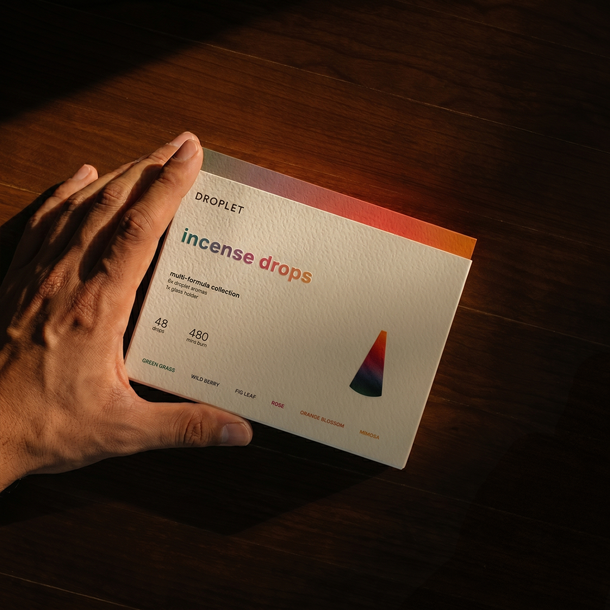 A hand holds a cream-colored card labeled 'DROPLET incense drops' over a dark wooden surface under dramatic warm lighting.