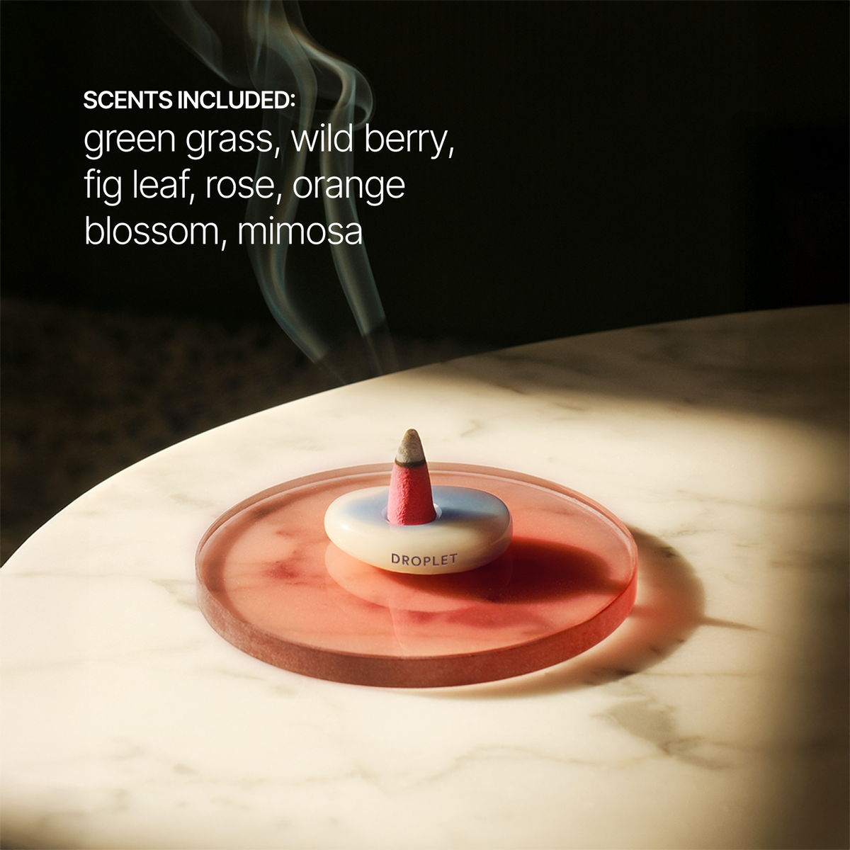 A droplet-style incense holder with a smoking incense stick sits on a translucent pink circular tray on a marble surface. Text on the image lists included scents: green grass, wild berry, fig leaf, rose, orange blossom, and mimosa.