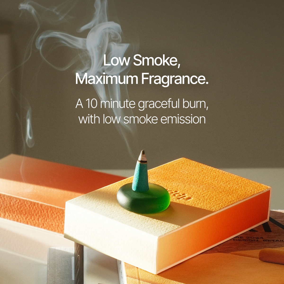 A green incense stick burning on a textured orange and cream book, with white smoke drifting upward. White text reads 'Low Smoke, Maximum Fragrance.