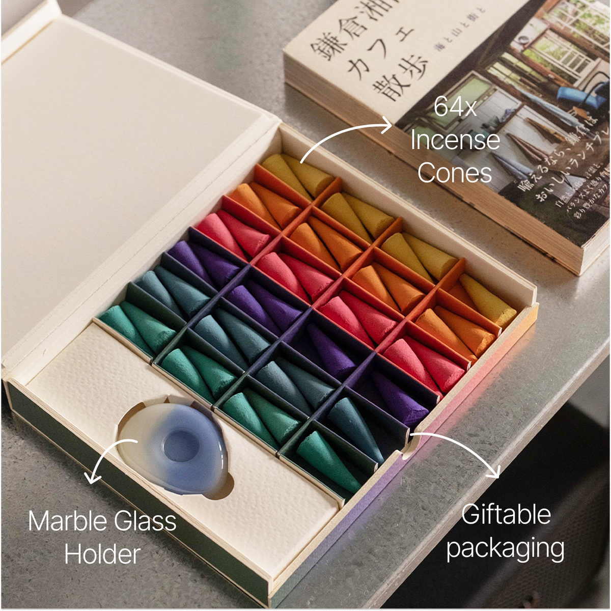 An open gift box containing a set of incense cones arranged in rows of colors, with a marble glass holder inside a slot. White text callouts read '64x Incense Cones,' 'Marble Glass Holder,' and 'Giftable packaging.