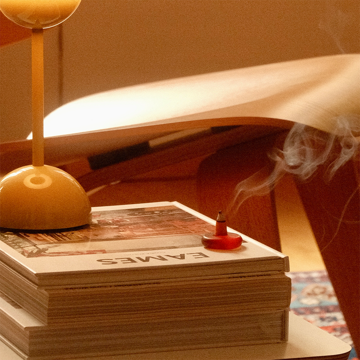 Stack of books with a smoking incense on top, next to a lamp, in a warm-toned room.