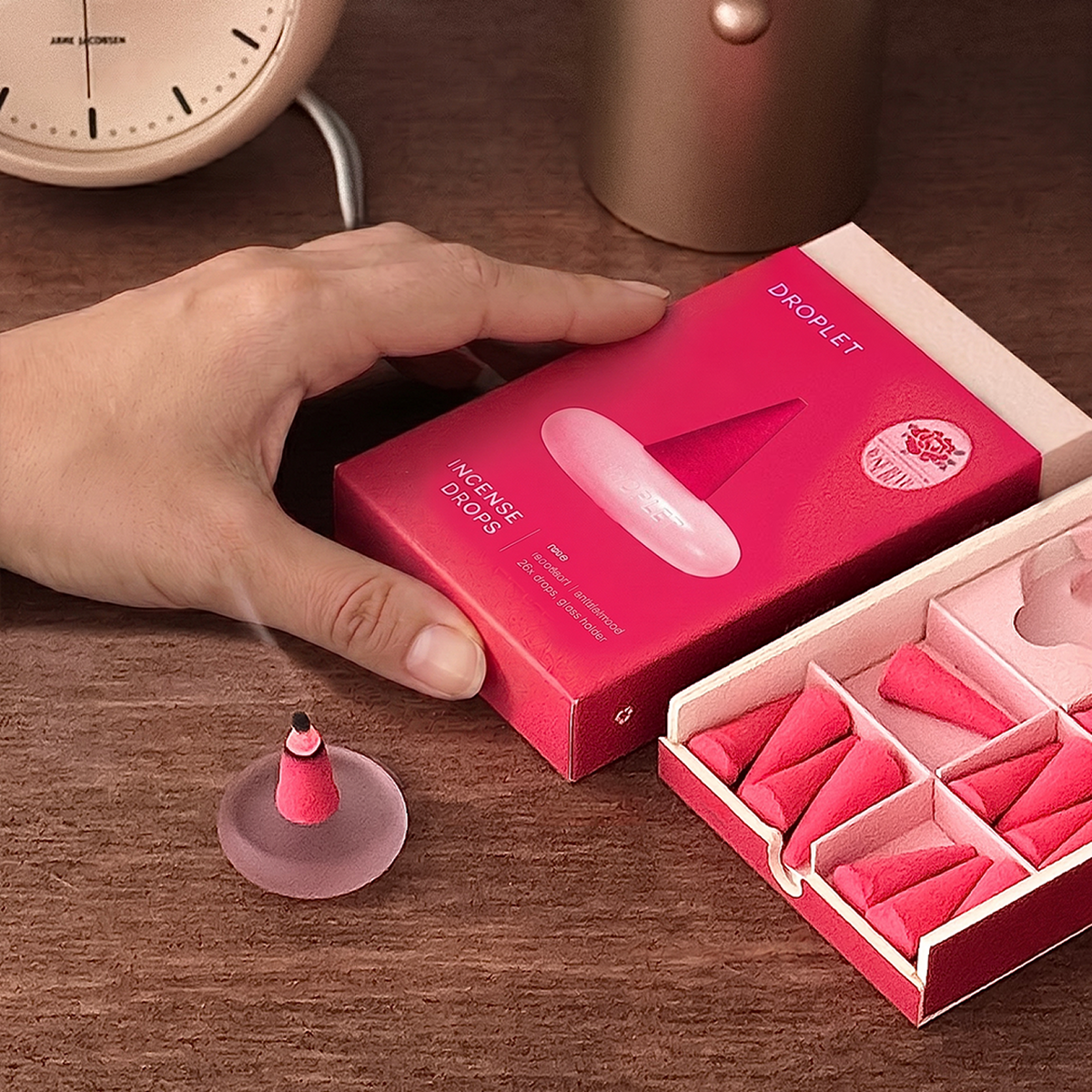 A hand holds a pink box labeled 'DROPLET INCENSE DROPS' next to an open tray of small pink incense cones on a wooden surface, with a blurred clock and metallic cup in the background.