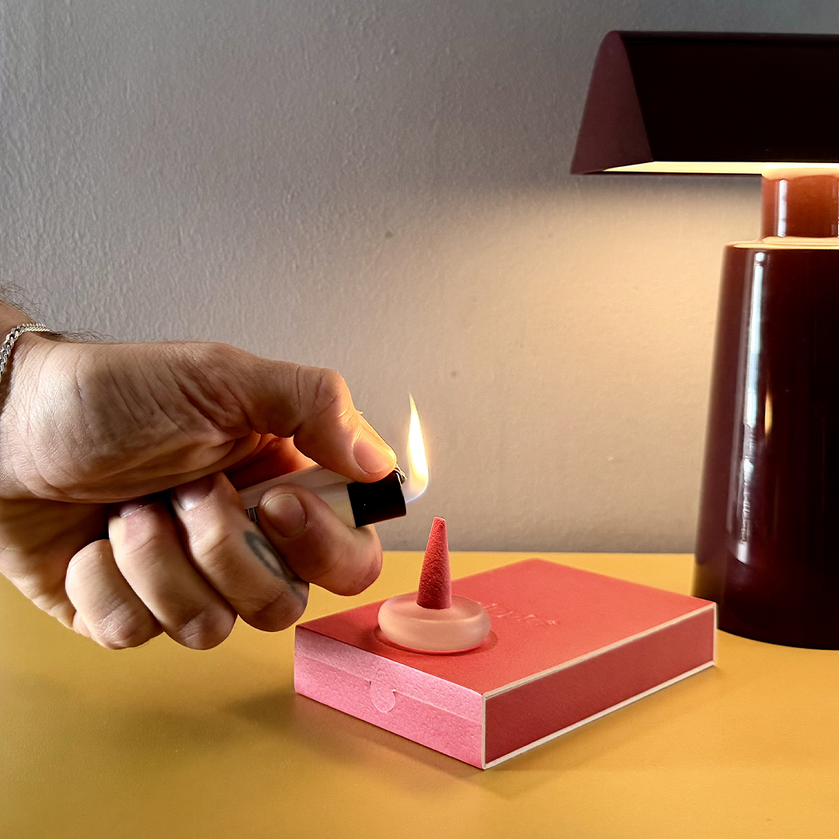 Hand lighting a small candle with a matchbox on a table next to a lamp.
