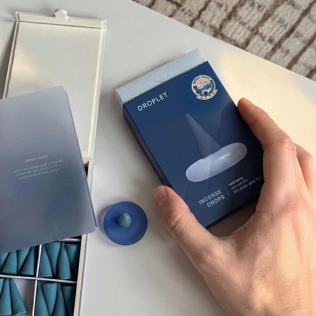 A hand holds a blue box of incense drops labeled 'DROPLET' and 'INCENSE DROPS' over an open tray containing multiple blue incense cones, on a white tabletop.