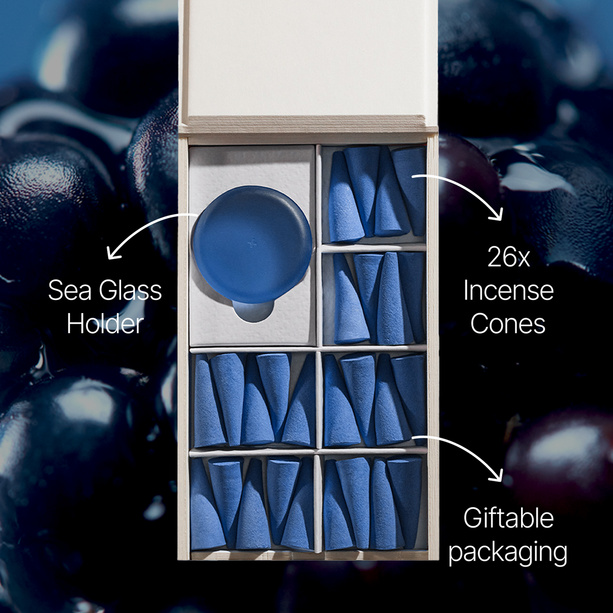 An open gift box containing a blue sea glass holder and 26 blue incense cones, shown with overlaid labels and arrows on a blurred dark berry background.