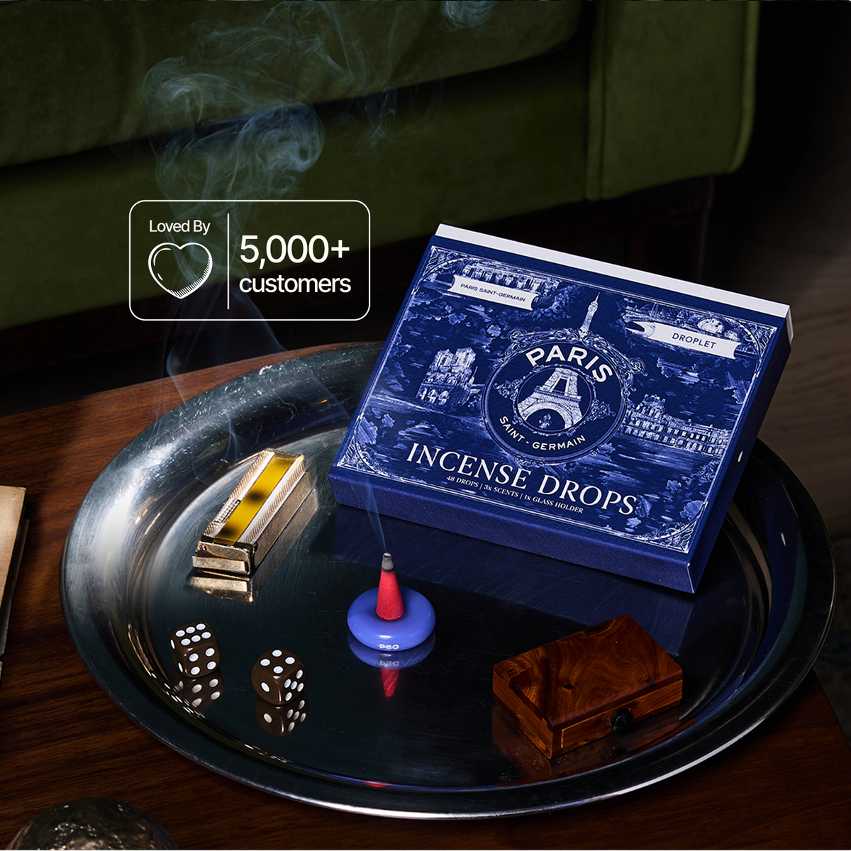 A blue Paris-themed box of Incense Drops sits on a dark tray with smoking incense, dice, and a small wooden box in a dim indoor setting. A badge reads 'Loved By 5,000+ customers'.
