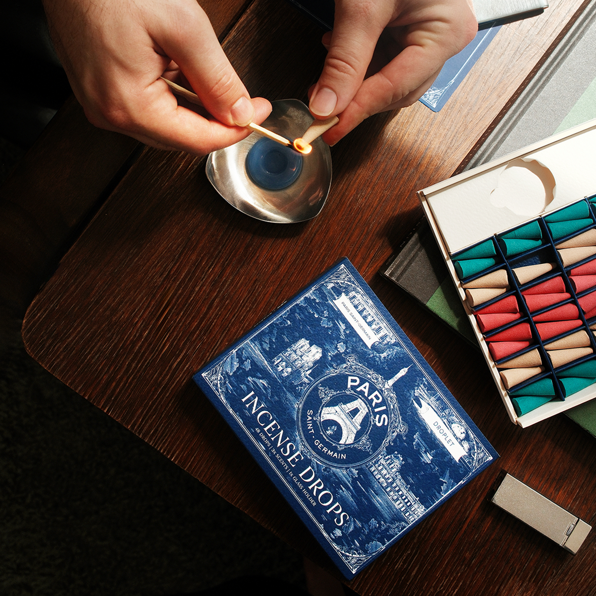 A person's hands light a match over a small metal dish on a dark wooden table, with a blue box of Paris Incense Drops and an open matchbox nearby.