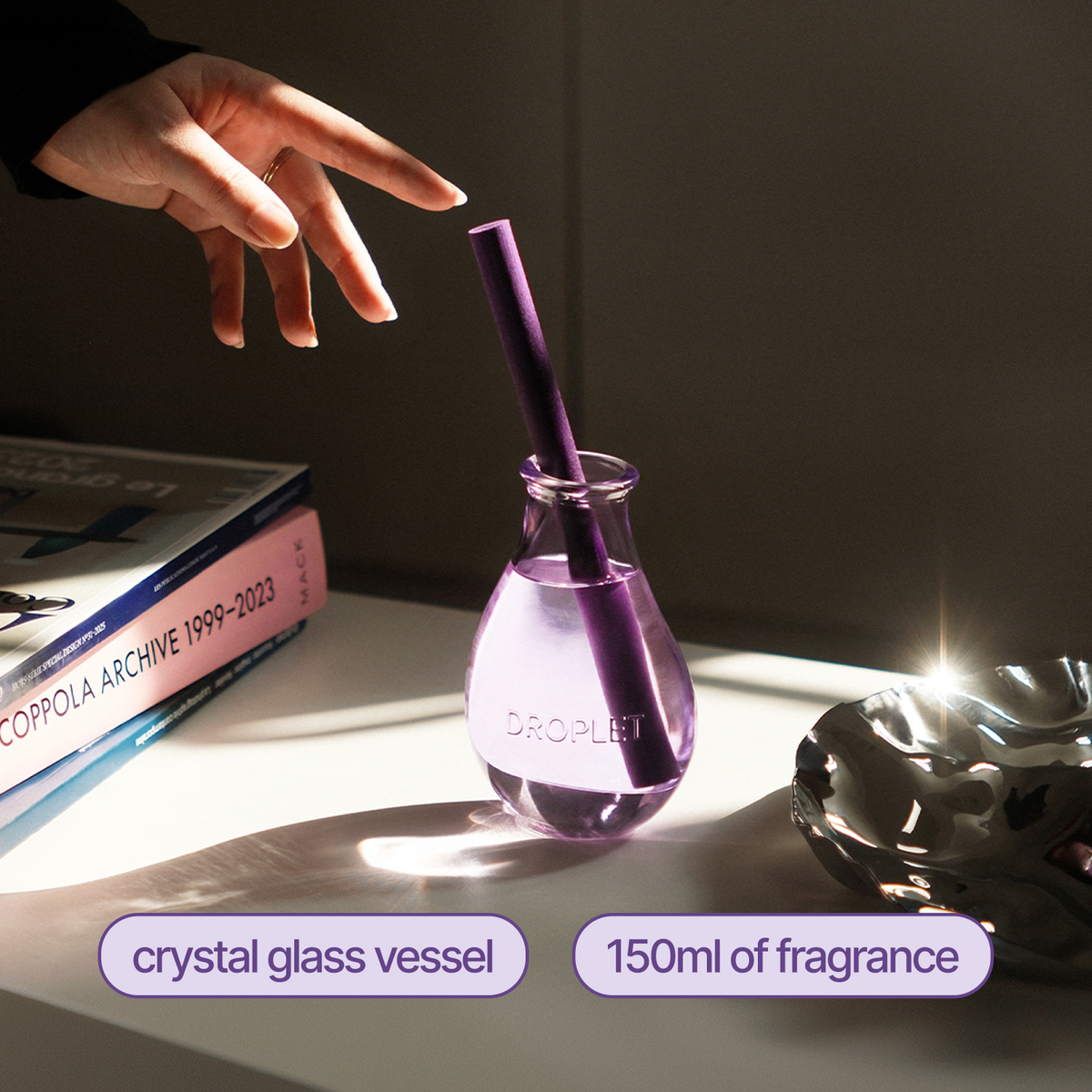 A purple droplet-shaped reed diffuser in a clear crystal glass vessel with a purple reed straw, shown on a table with a hand reaching toward it. Text labels identify the crystal glass vessel and 150ml fragrance.