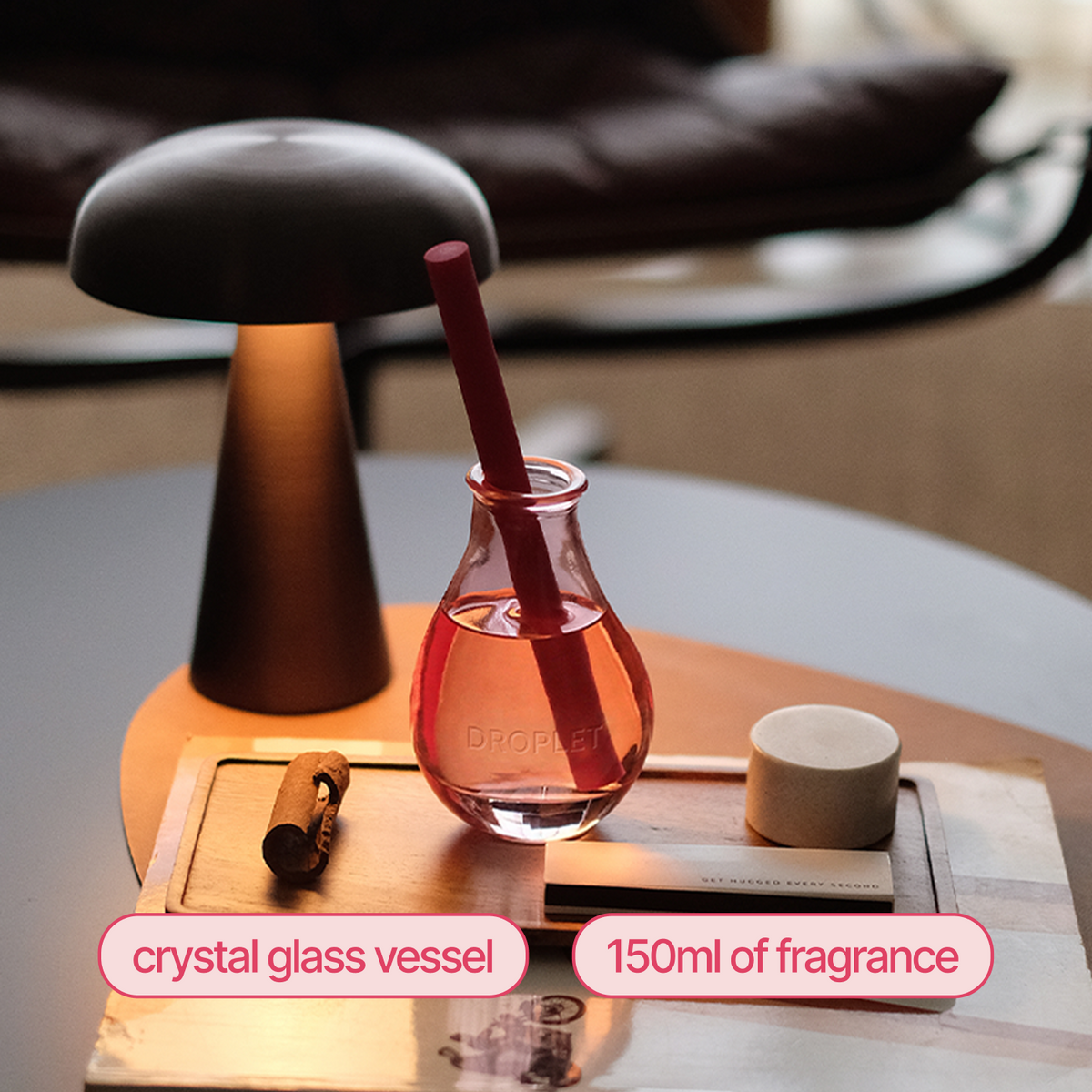A crystal glass diffuser vessel filled with pink fragrance and a dark red straw, shown on a table beside a lit mushroom-shaped lamp, a small candle, and a cork. Text labels identify the crystal glass vessel and 150ml of fragrance.