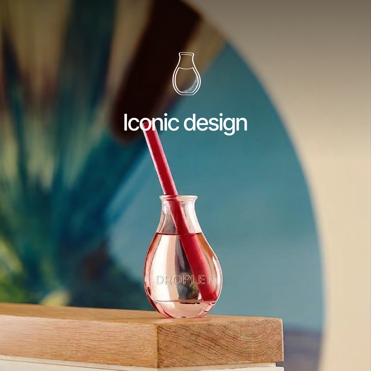 A small glass perfume bottle with a red cylindrical diffuser inside, placed on a wooden surface against a softly blurred blue and beige background. White text reads 'Iconic design' and the bottle has faintly visible 'DROPLET' branding.