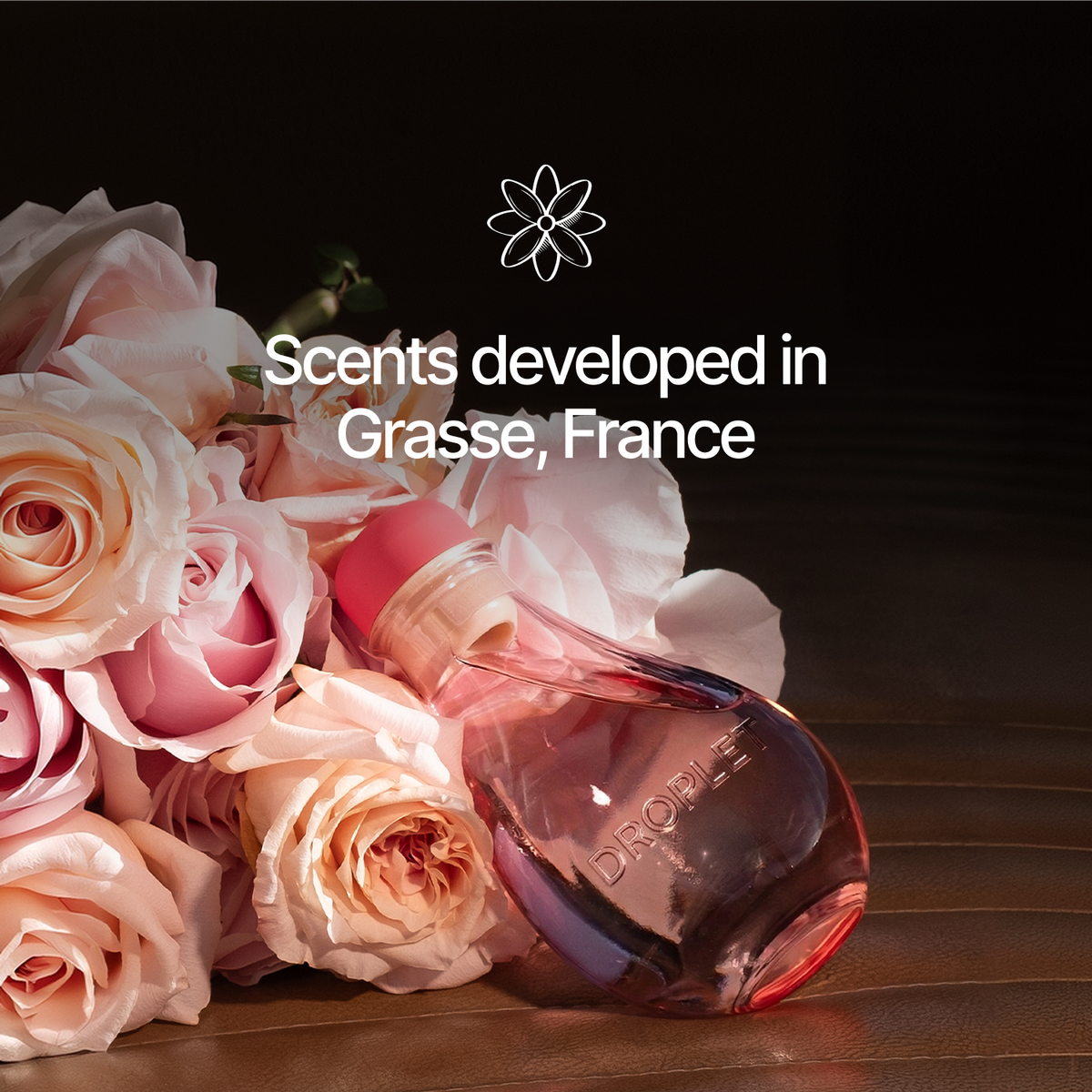 A perfume bottle with a pink cap and translucent pink glass lies on a wooden surface beside a bouquet of pale pink roses. White text reads 'Scents developed in Grasse, France' with a flower icon above, against a dark background.