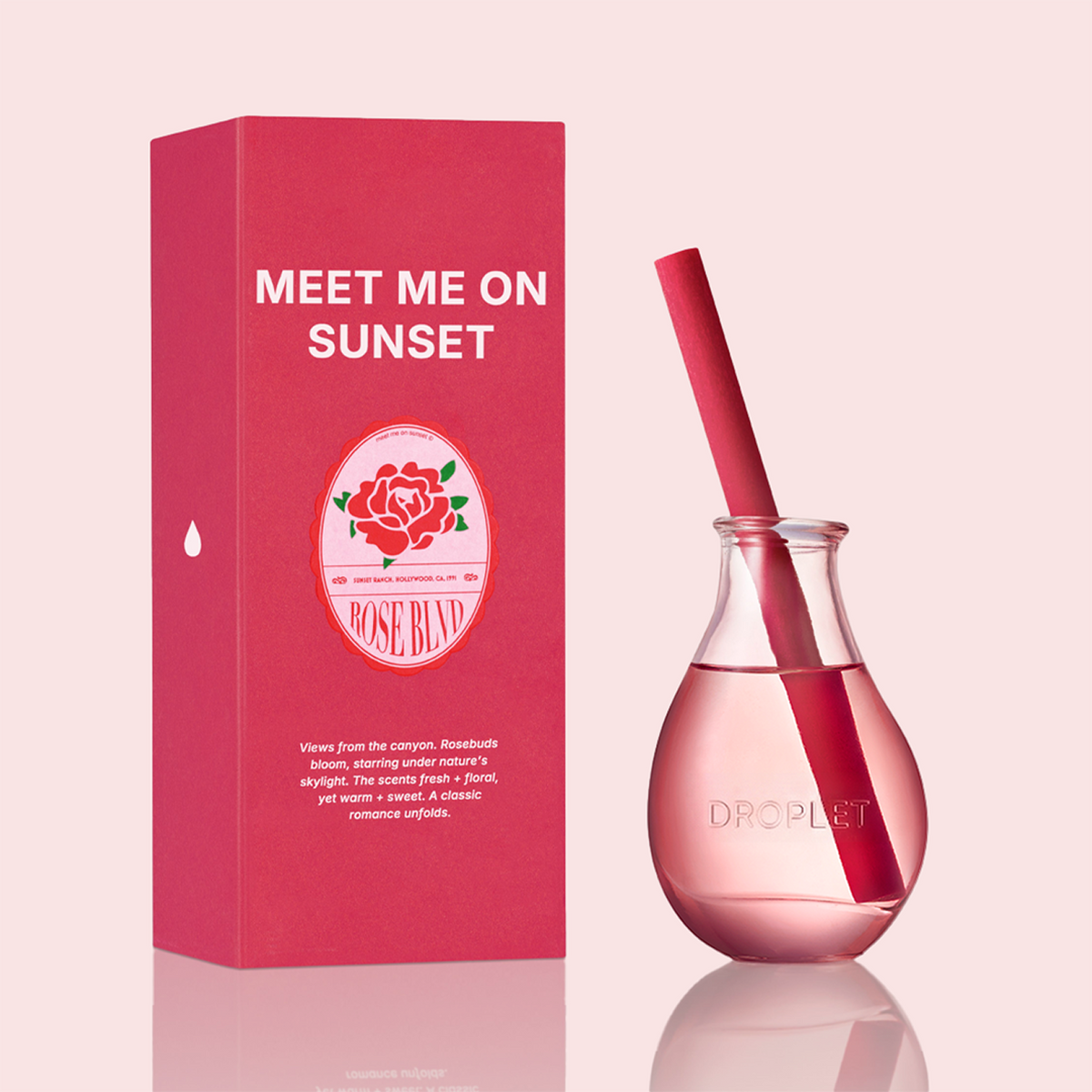 Droplet Rose Bud 'Meet Me On Sunset' fragrance set featuring a pink rectangular box with white text and a clear droplet bottle filled with pink liquid and a pink straw, on a pale pink background.