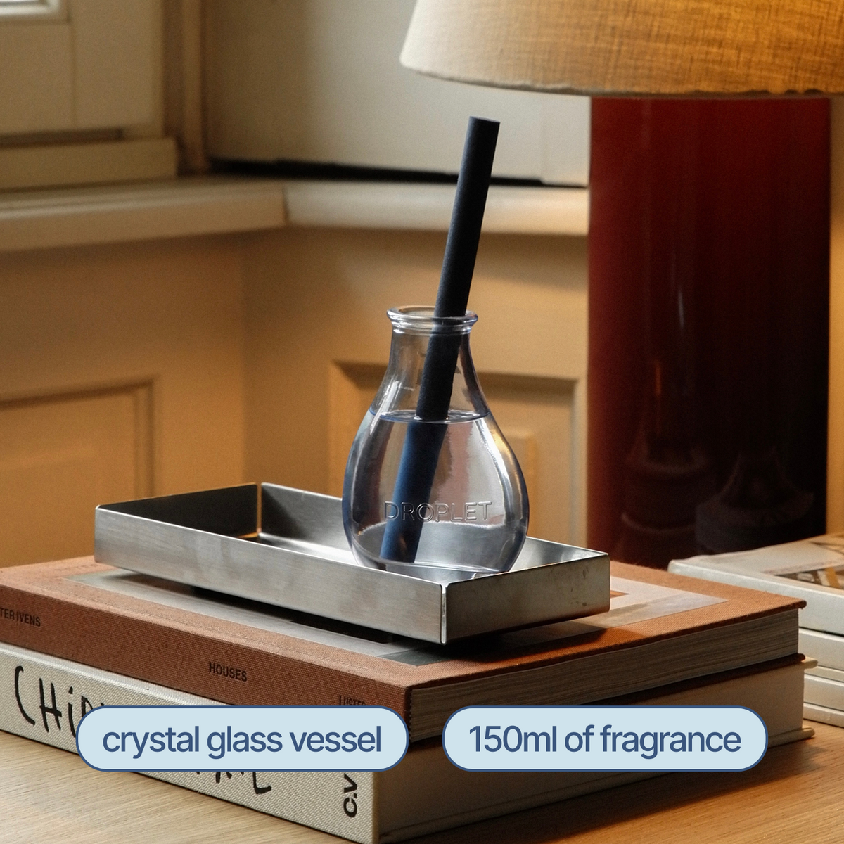 A clear crystal glass fragrance vessel with a black straw sits on a rectangular metal tray, placed on top of stacked books in a warm indoor setting. Text labels identify the crystal glass vessel and 150ml of fragrance.