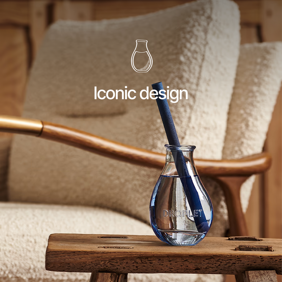 A clear glass droplet-shaped vase with a blue textured straw sits on a wooden table in front of a cozy beige armchair. White text above reads 'Iconic design' with a simple vase outline.