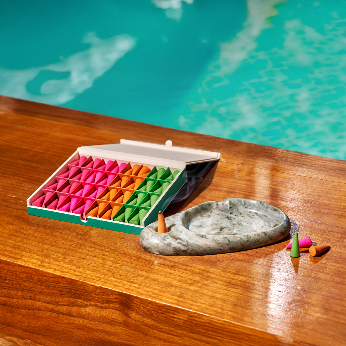 A set of colorful triangular desk accessories arranged in a green and white box on a wooden table, with a gray stone tray and a few loose pieces in front. Turquoise water is visible in the background.