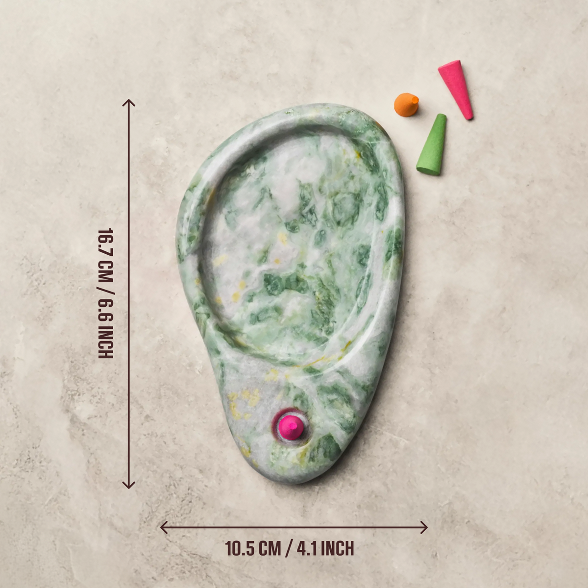 A carved stone ear-shaped object with green marbling and a small pink gemstone, shown on a beige textured background with measurement arrows and text indicating 16.7 cm height and 10.