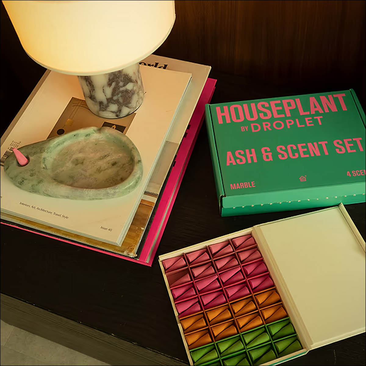 A styled product shot of the Houseplant by Droplet Ash & Scent Set, showing a green product box, an open tray of colorful marble scent cards, and a marble ashtray resting on books next to a lamp.