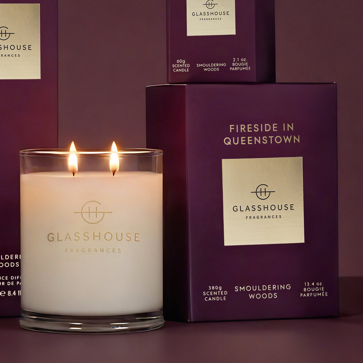 A lit double-wick scented candle in a clear glass jar with visible flames, placed in front of matching purple packaging boxes. The candle and boxes are branded GLASSHOUSE FRAGRANCES, with the scent name Fireside in Queenstown visible on the front.