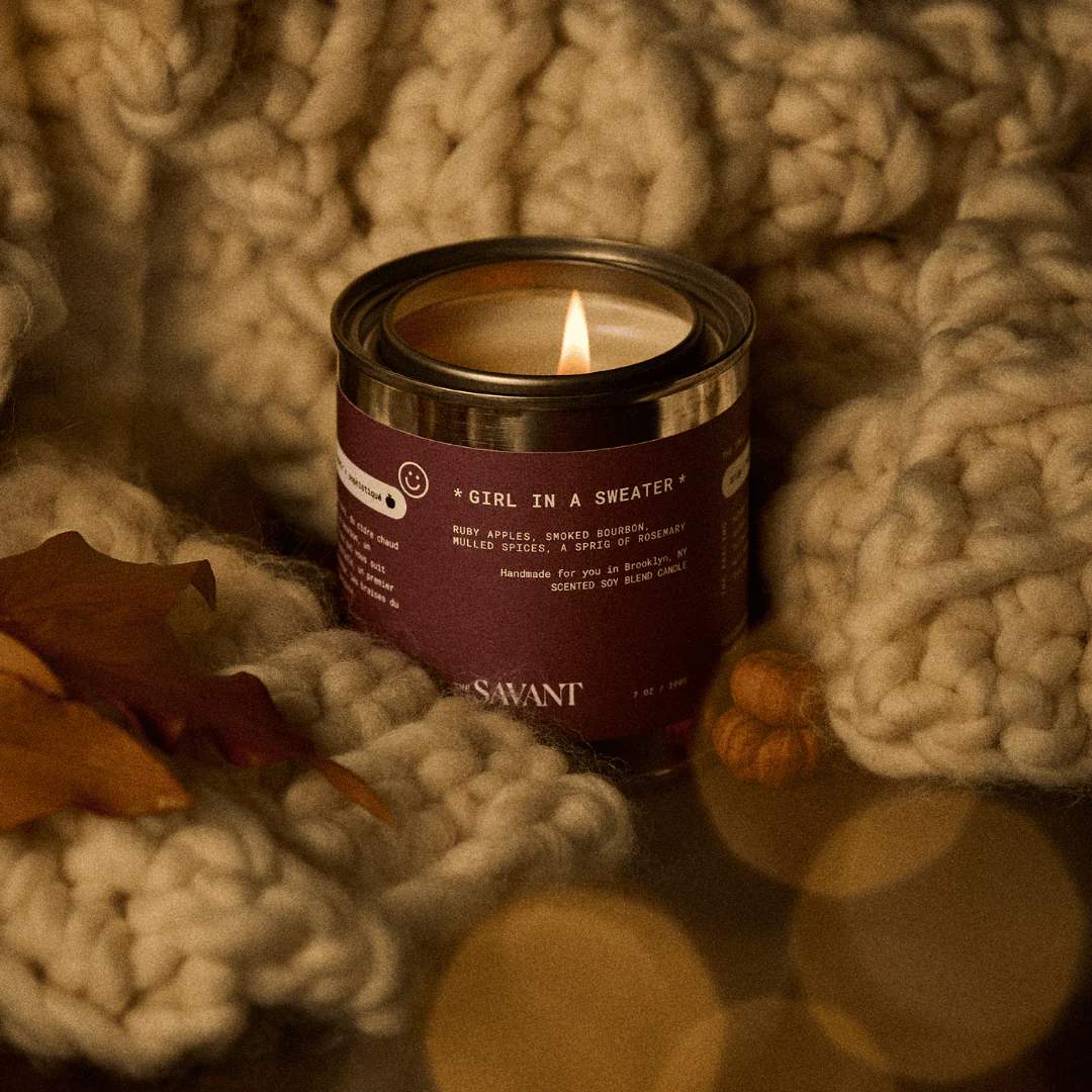 Candle in a metal tin labeled 'Girl in a Sweater' by The New Savant, surrounded by textured fabric and leaves.