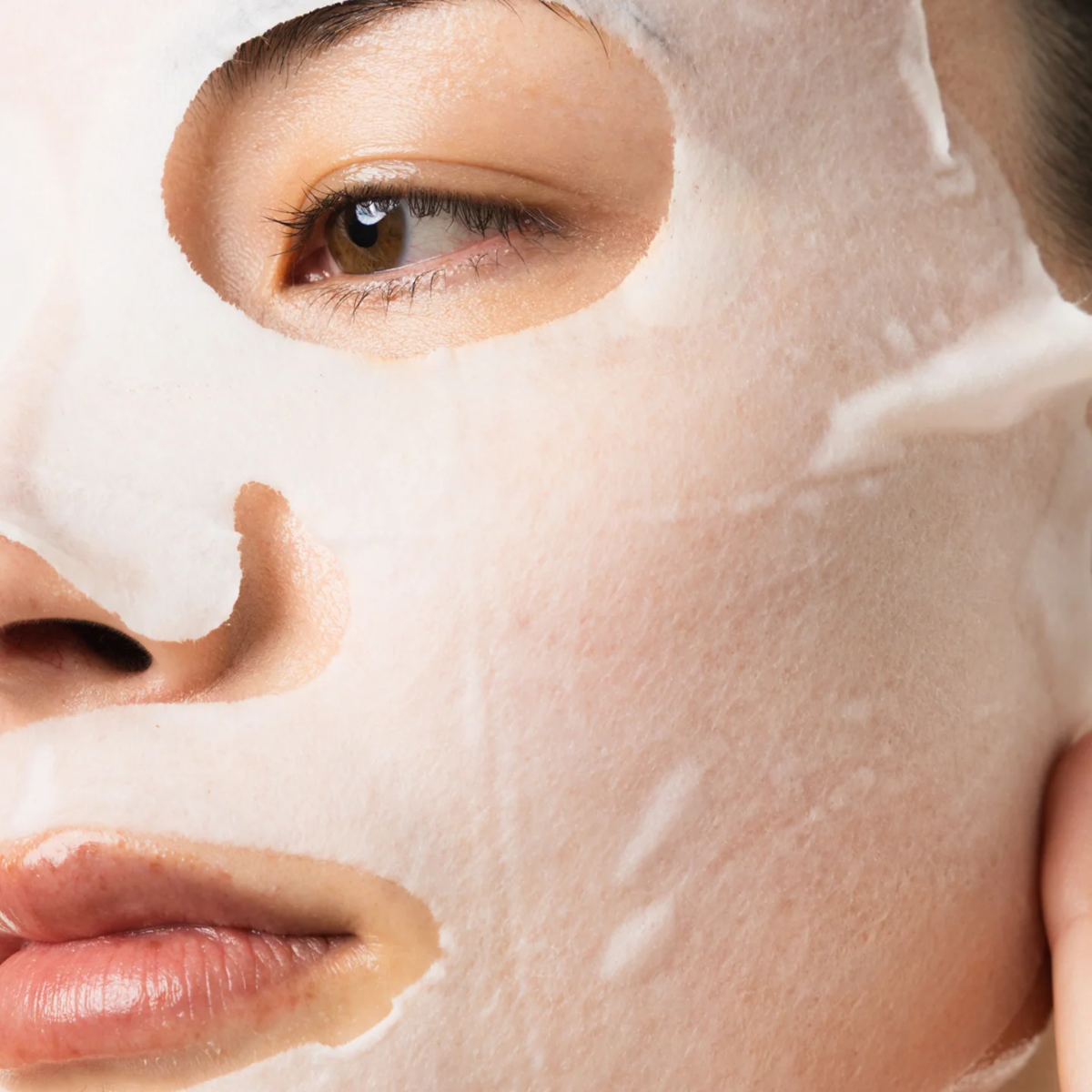 Close-up of a person wearing a translucent white sheet mask over part of their face, with the eye and lips visible.