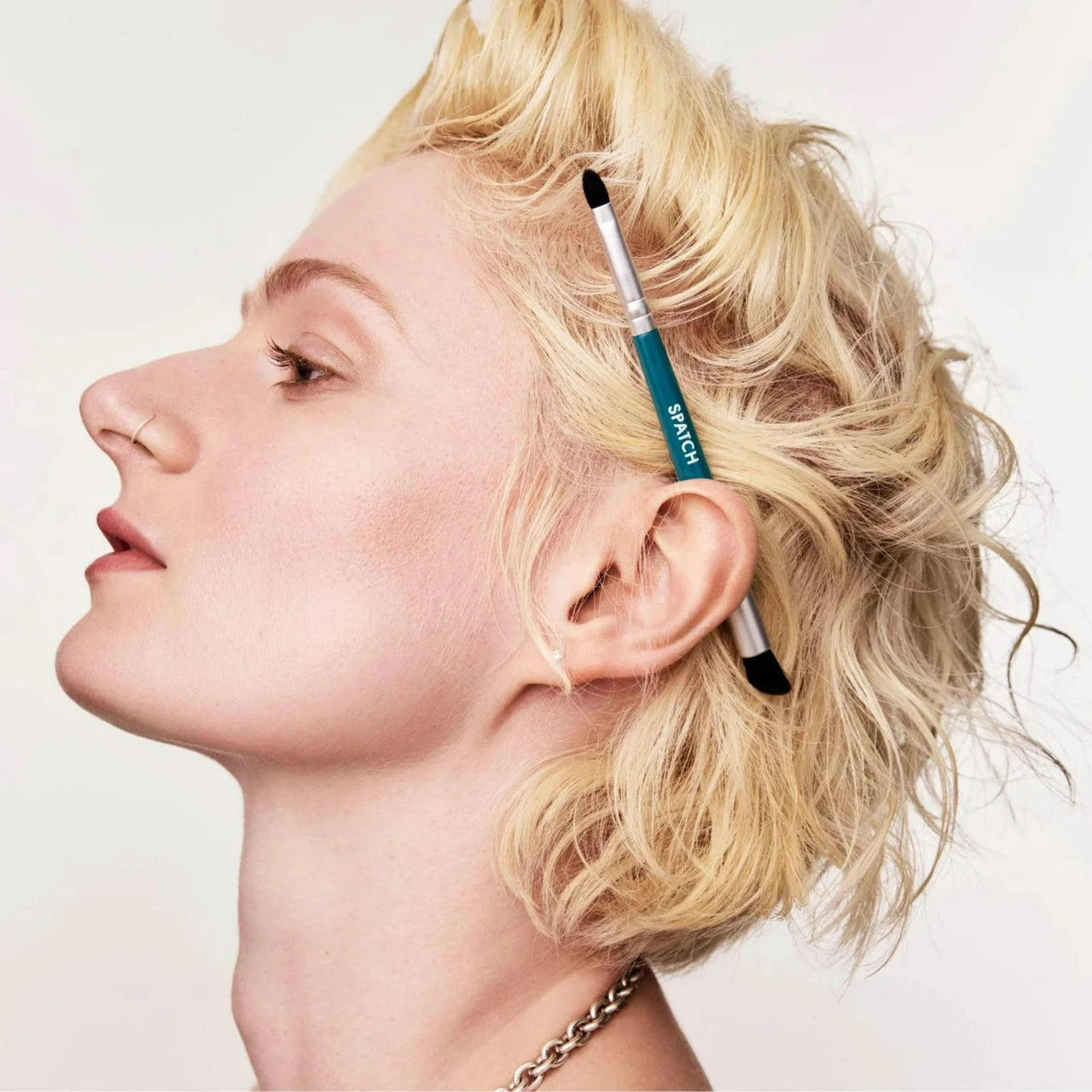 Woman with blonde hair and a makeup brush near her ear on a light background