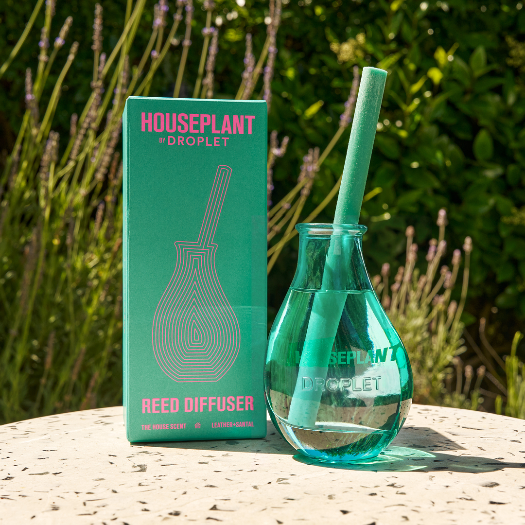 A teal-green reed diffuser box and matching glass bottle with a reed diffuser straw, placed on a stone table outdoors with blurred lavender and greenery in the background.