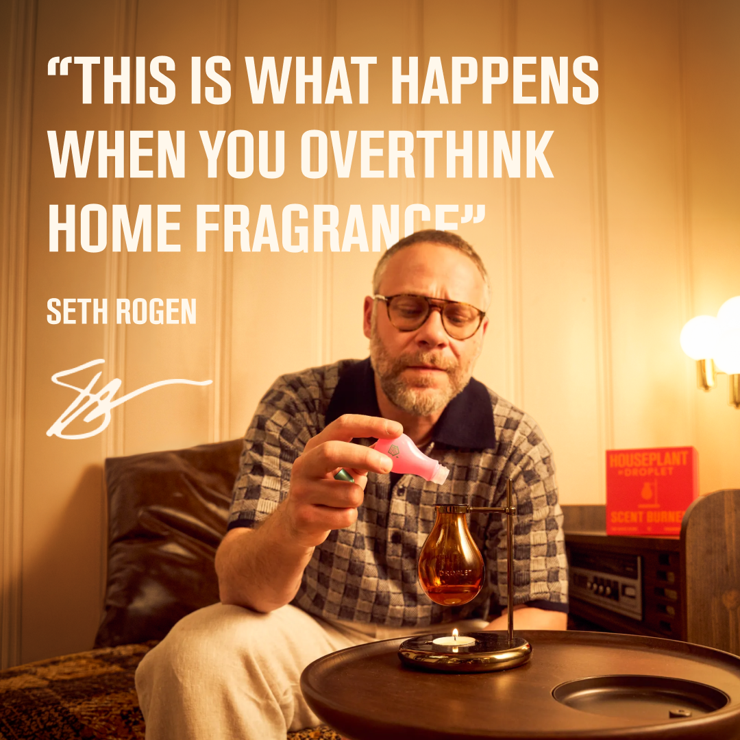 Seth Rogen in a checkered shirt and glasses sits in a warmly lit room, holding a pink diffuser above a small amber glass bottle on a round table. Large white text at the top reads, "THIS IS WHAT HAPPENS WHEN YOU OVERTHINK HOME FRAGRANCE" with Seth Rogen's name and signature below.