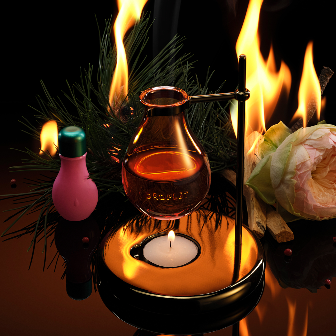 A glass perfume distillation apparatus with amber liquid in a bulb, a small pink bottle with a green cap, pine branches, wood, and flames against a black background.