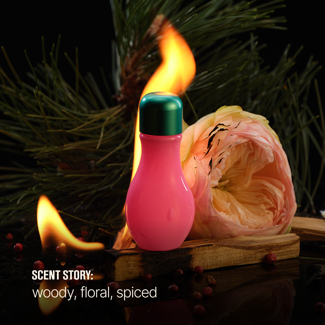 A pink perfume bottle with a metallic green cap stands on a wooden board with pine branches, small red berries, and flames in the background. Text at the bottom left reads 'SCENT STORY: woody, floral, spiced'.