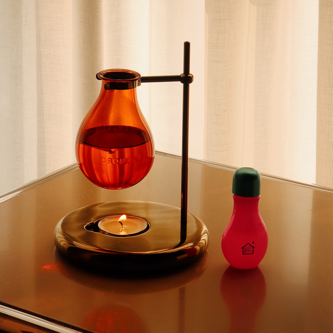 An amber glass heated vessel suspended in a magnetic levitation setup on a brass stand, with a small lit candle in a matching brass base and a pink bottle with a green cap beside it, set on a reflective table in front of sheer curtains.
