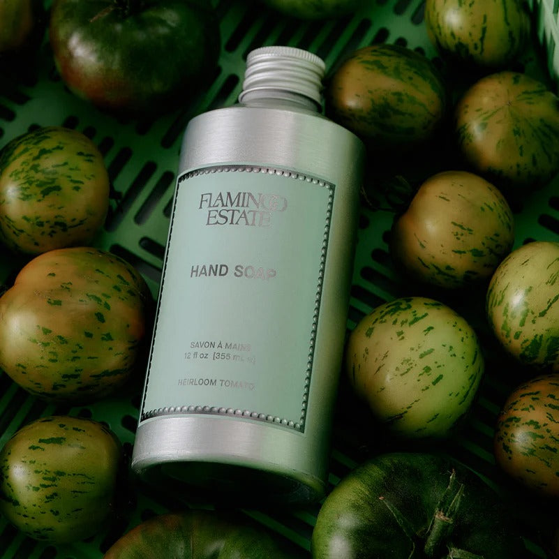 Hand soap bottle labeled 'Flamingo Estate' surrounded by green tomatoes on a green basket.