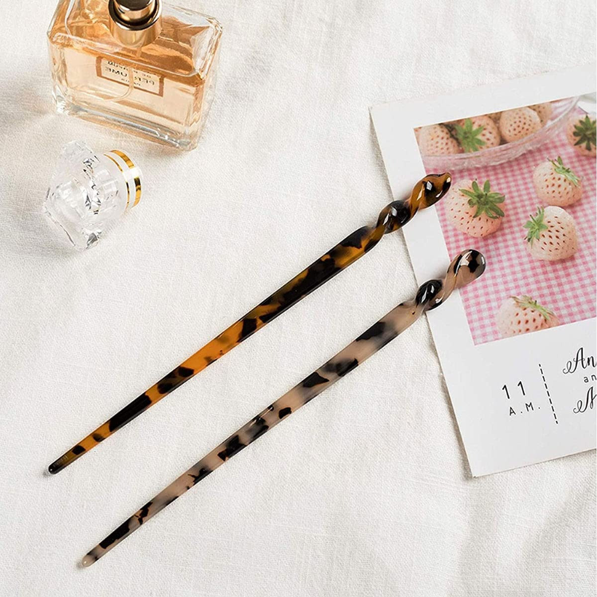 Two tortoiseshell hair sticks on a white surface with a small bottle of perfume and a card with strawberries in the background.