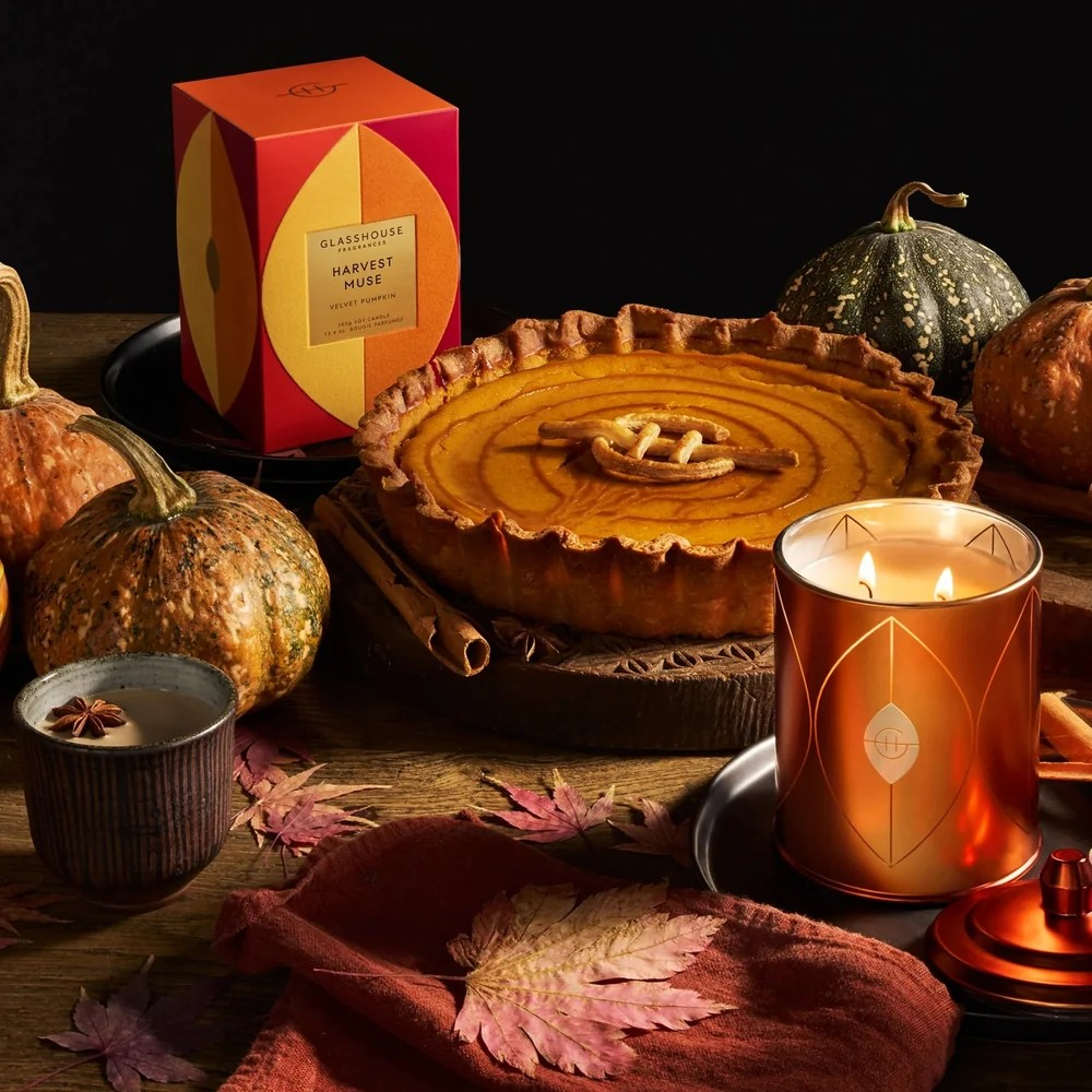 A styled autumn tabletop scene featuring a glasshouse scented candle in a metallic copper-toned tin next to a pumpkin pie, with pumpkins, leaves, a cup, and the product box in the background.