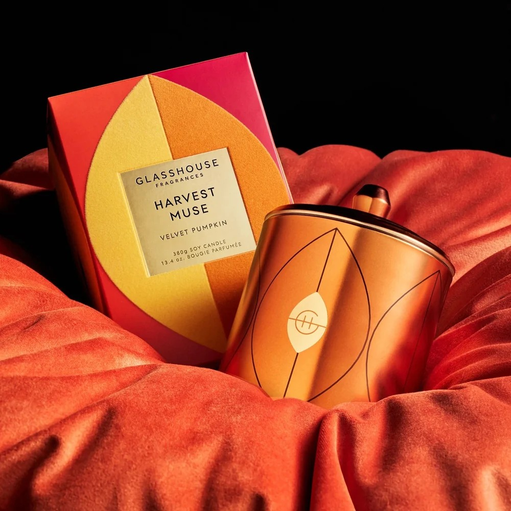 A GLASSHOUSE Fragrances Harvest Muse candle in a metallic copper-orange container with a leaf design, displayed next to its colorful red, orange, and yellow box on draped velvet fabric.