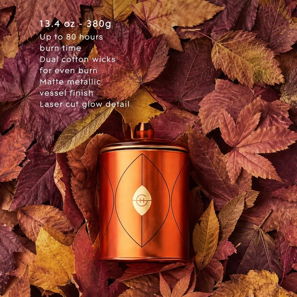 An orange metallic cylindrical candle tin with a geometric line design sits on a bed of dried autumn leaves. White text on the upper left lists product features including 13.