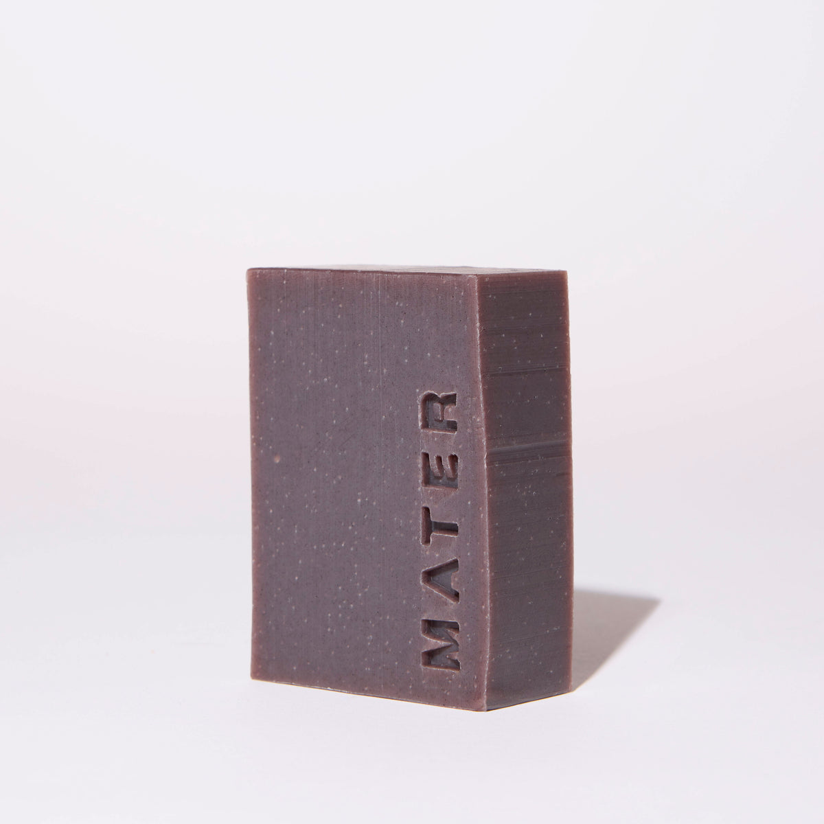 A rectangular bar of MATER soap in a dark plum-brown color, standing upright on a clean white background with a soft shadow cast to the right.