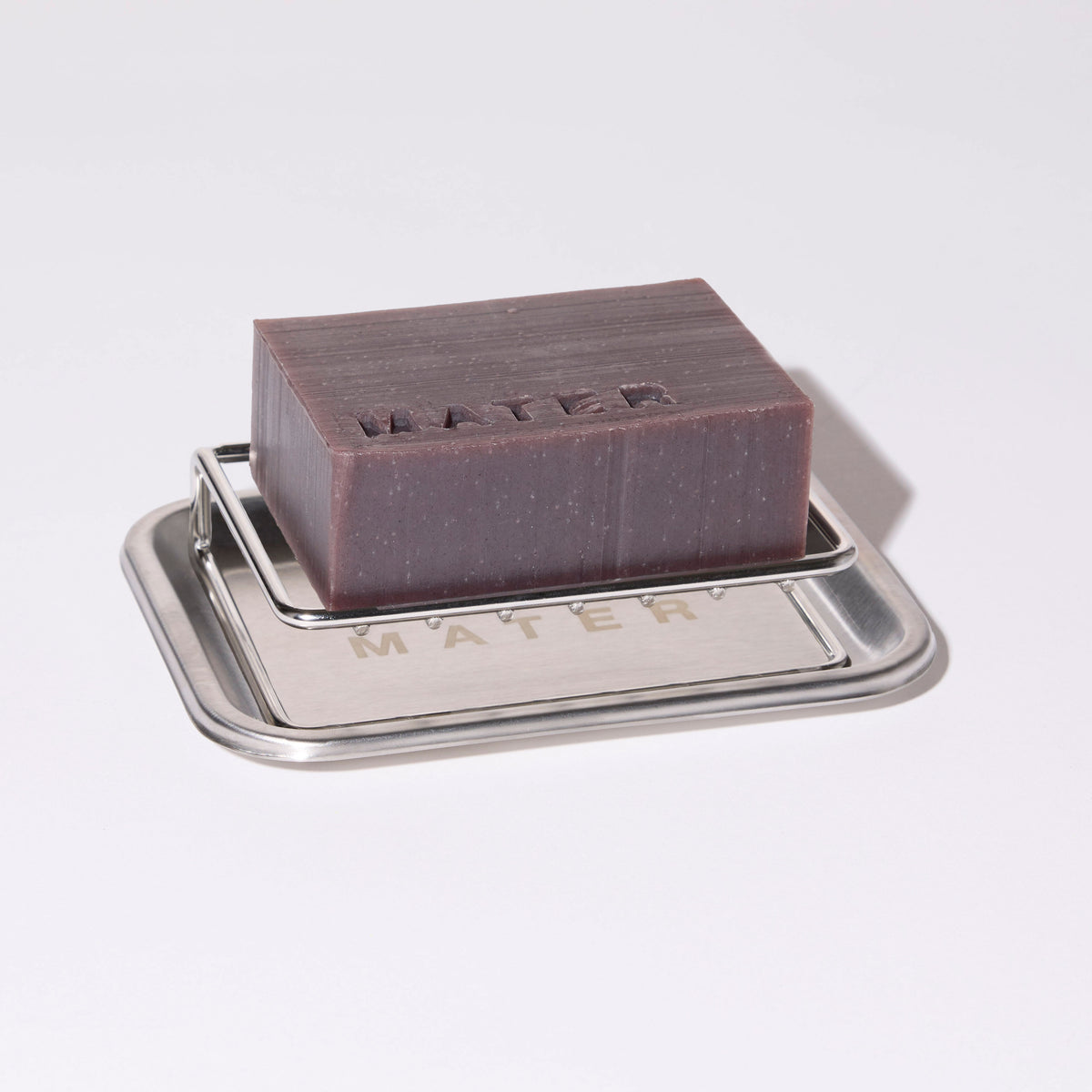 A rectangular dark brown soap bar embossed with the word 'MATER' sits on a small silver metal tray with 'MATER' also embossed on the front, photographed on a clean white background.