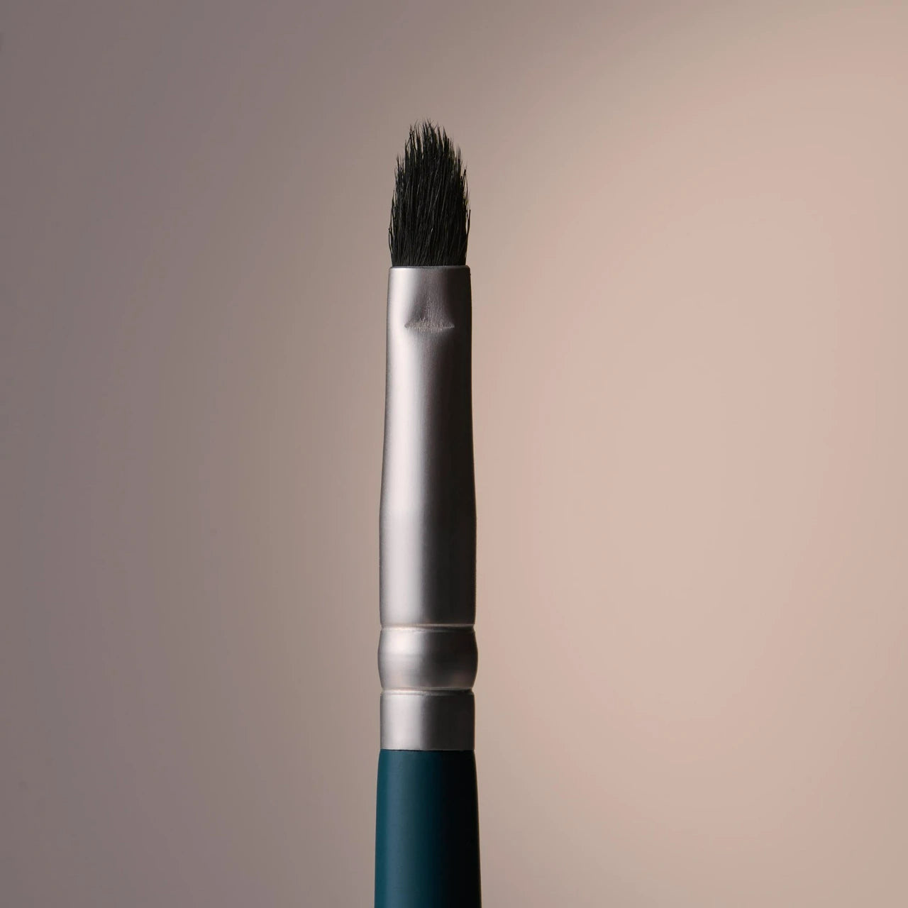 A close-up studio shot of makeup brush with dark bristles, a silver ferrule, and a dark teal handle against a soft beige gradient background.