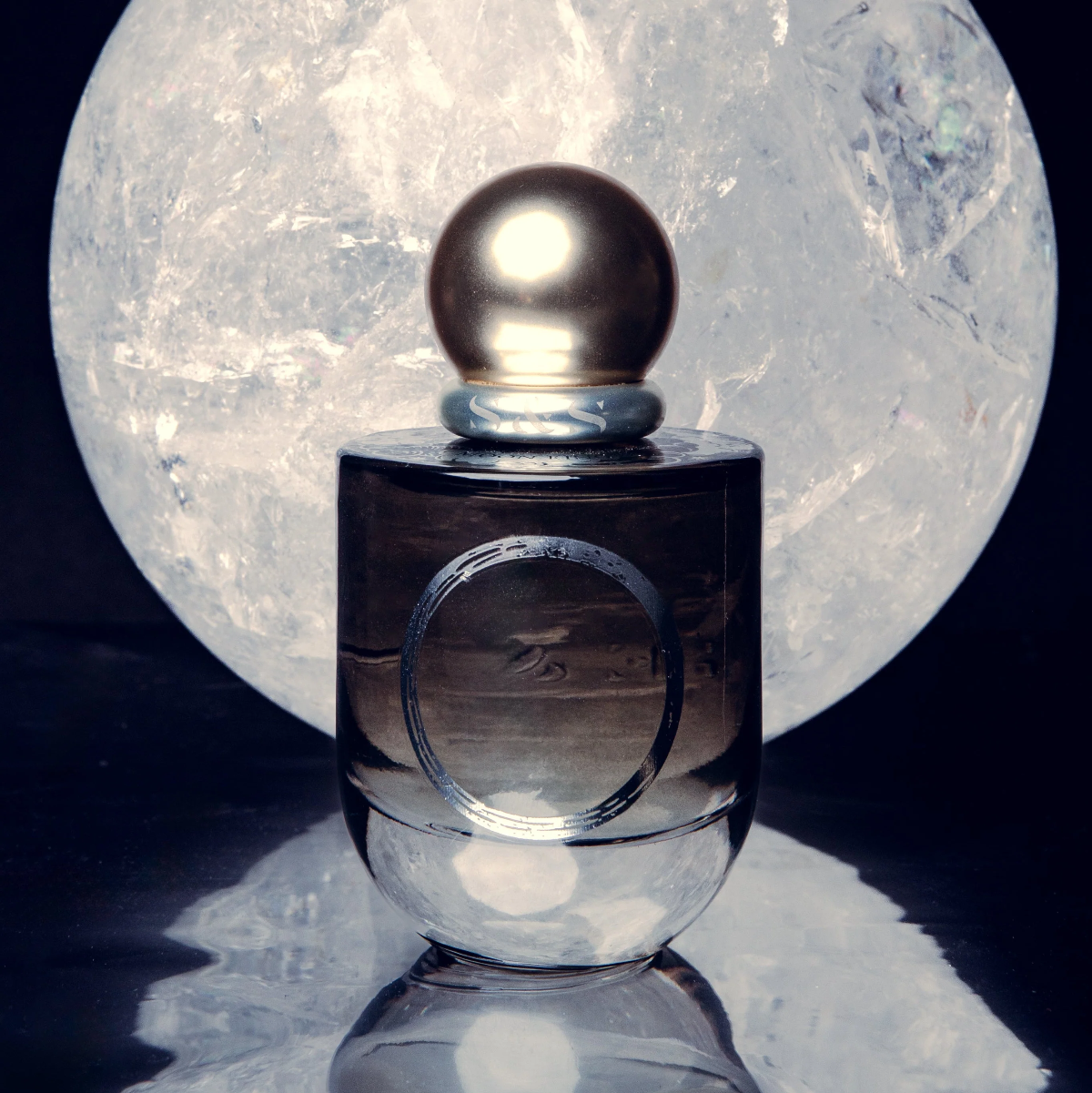 A dark amber glass perfume bottle with a metallic gold spherical cap, set against a large glowing moon backdrop on a reflective black surface.