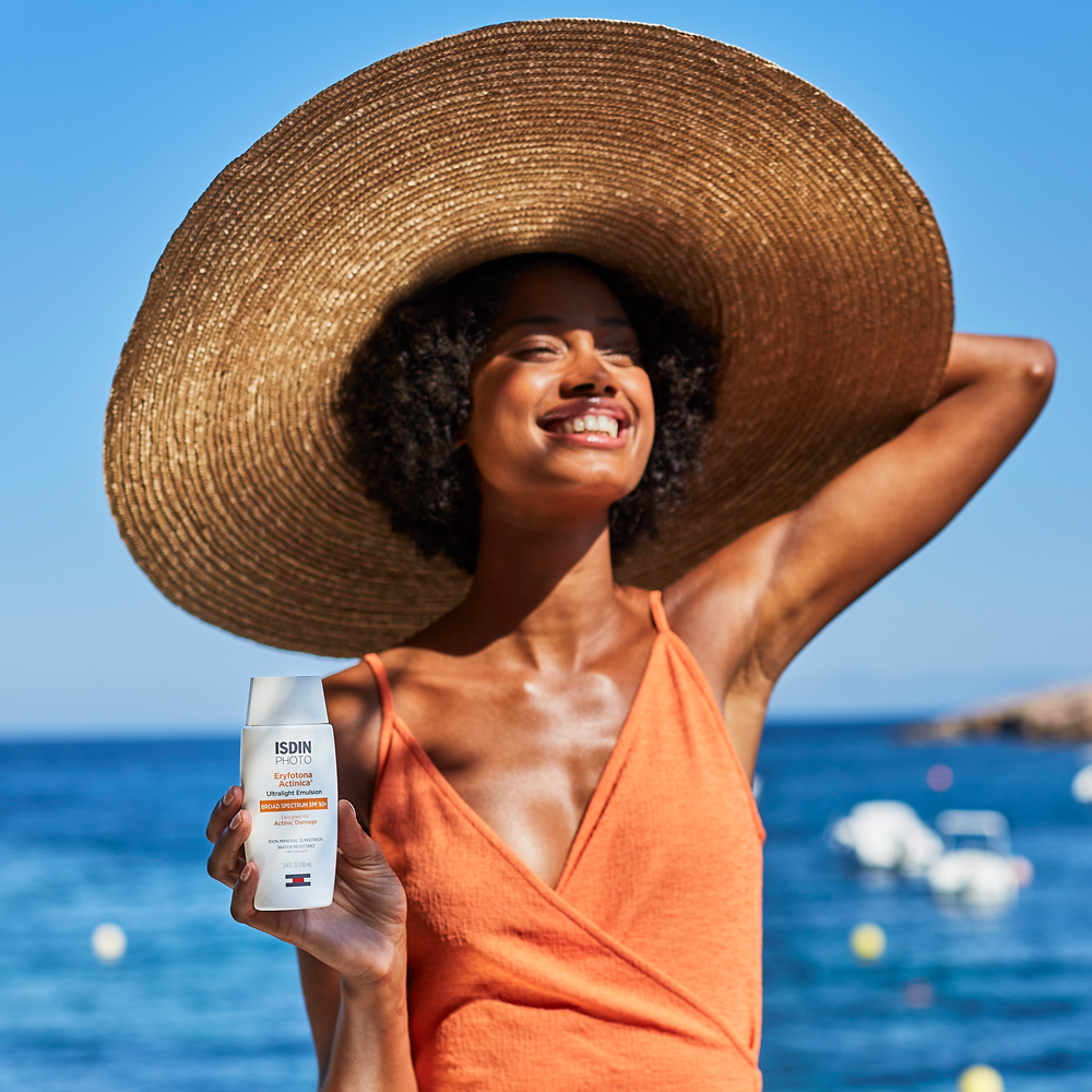 A woman wearing a large straw sun hat and an orange swimsuit stands by the ocean holding ISDIN Eryfotona Actinica SPF 50+ bottle