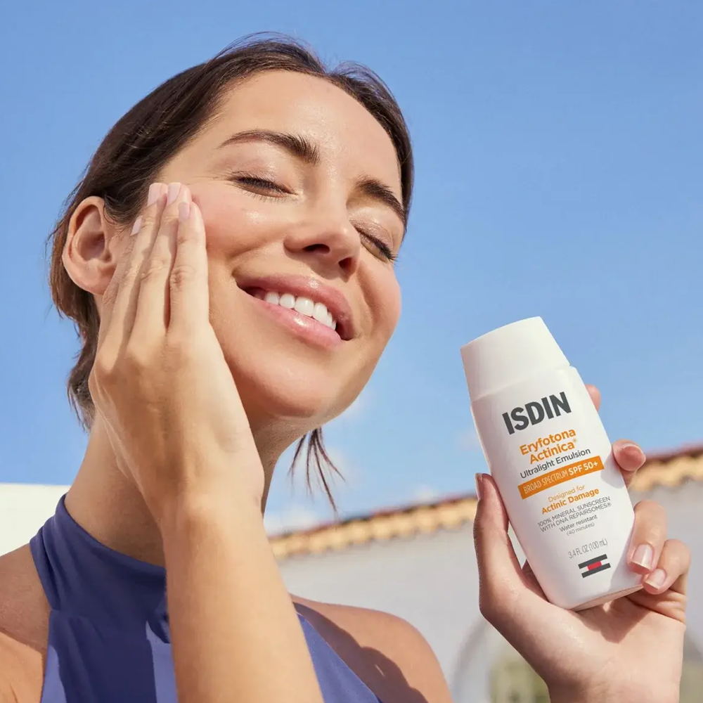 A woman outdoors under a blue sky applies ISDIN Eryfotona Actinica SPF 50+ to her face while holding the bottle