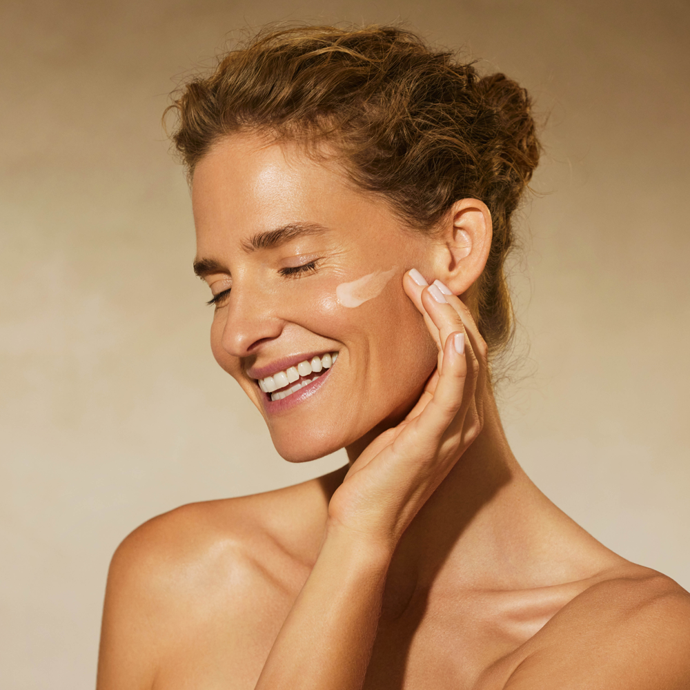 A close-up of a smiling woman with her eyes closed, applying ISDINCEUTICS Melaclear Advanced 3% product on her cheek against a warm beige background.